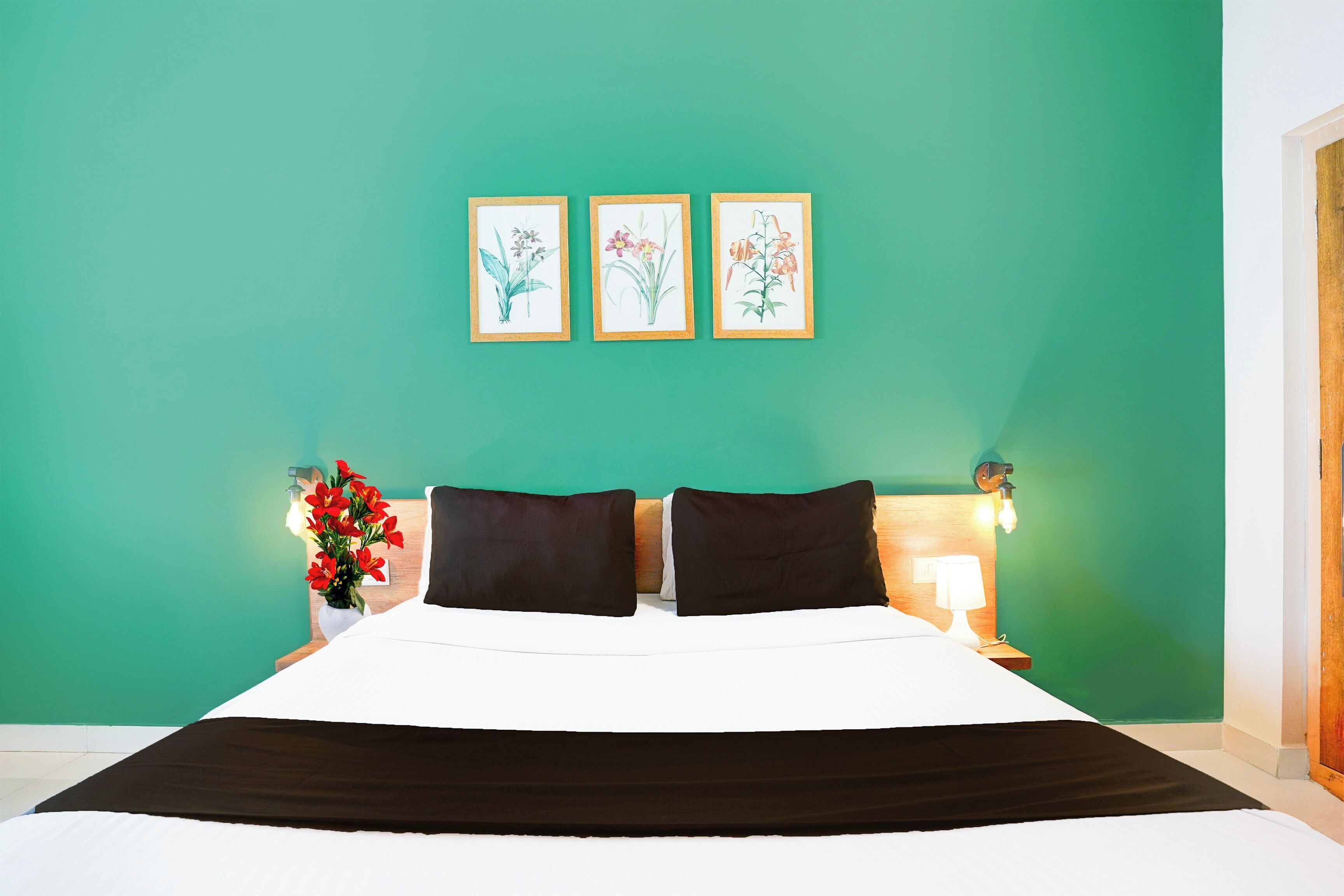 Habitación de hotel con una cama, dos almohadas, una mesita de noche con una lámpara y tres ilustraciones botánicas enmarcadas en la pared.
