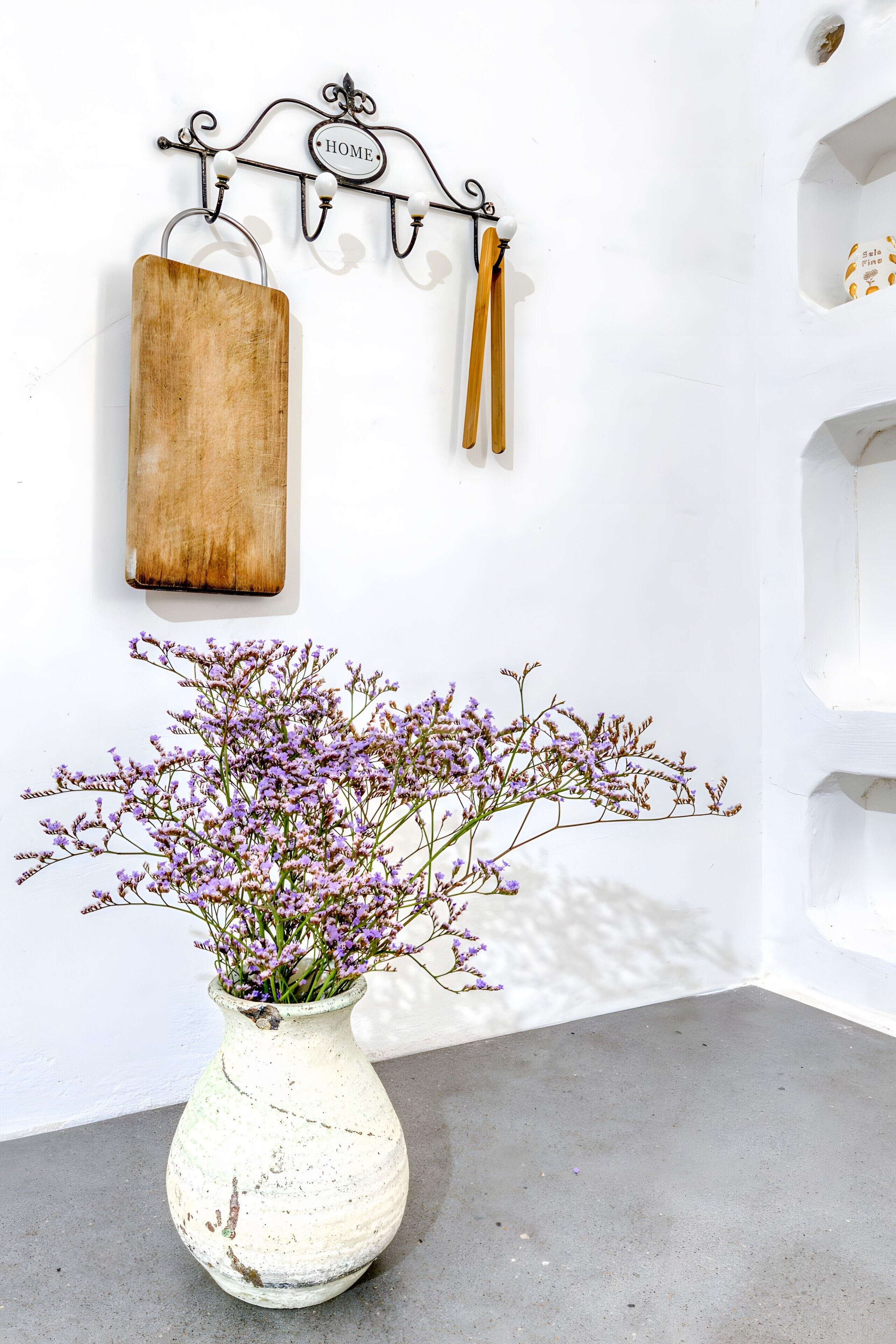 Eine weiße Wand mit einem rustikalen Holzschneidebrett, einem Metallhakenregal und einer weißen Vase mit violetten Blumen.