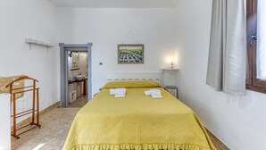 Une chambre à coucher avec un lit, une table de chevet, une commode, une fenêtre avec des rideaux, un tableau accroché au mur et une salle de bain visible à travers une porte ouverte.