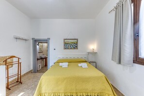Un dormitorio con cama, mesita de noche, cortinas en la ventana, un cuadro en la pared y un baño visible a través de una puerta abierta.