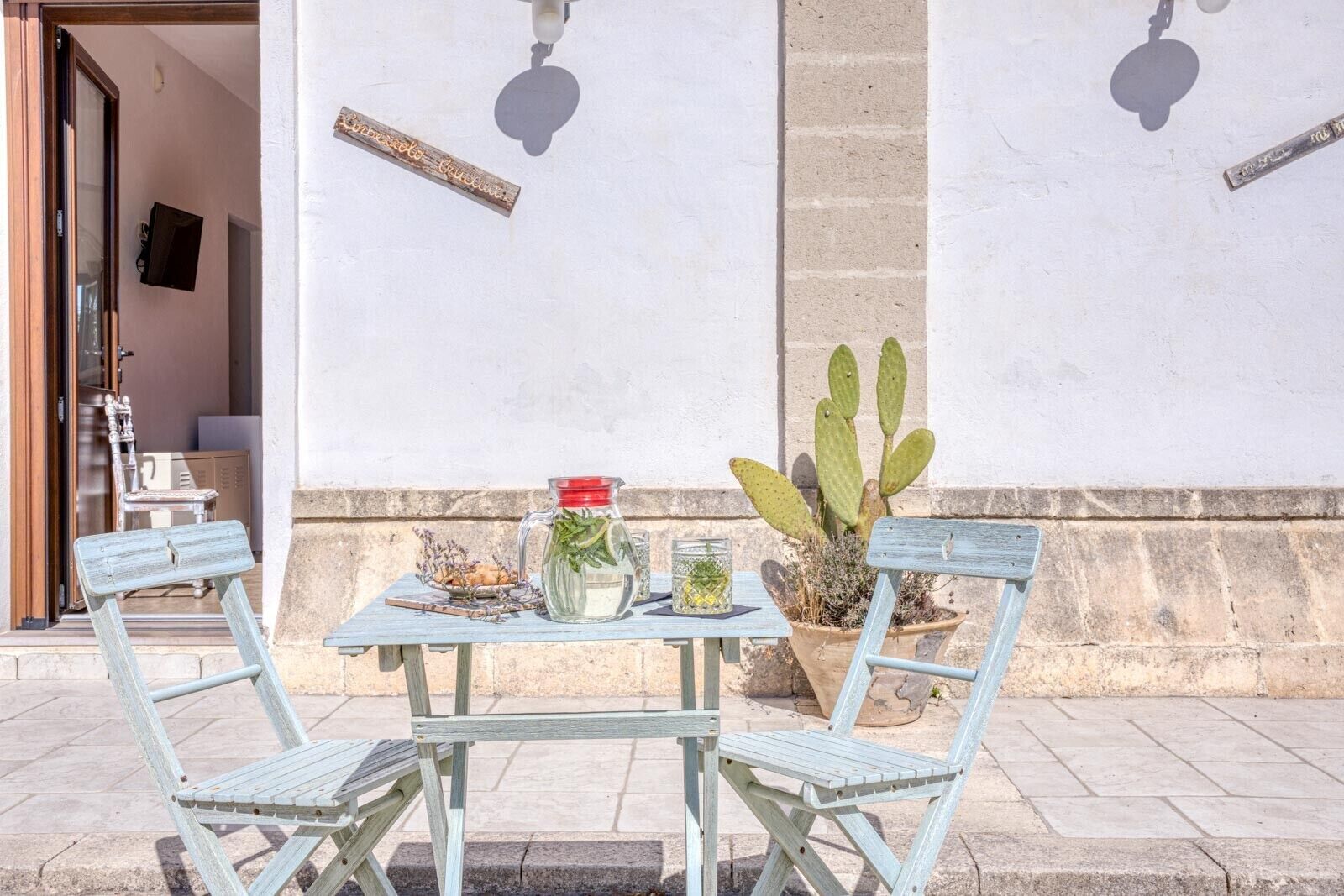 An outdoor seating area with a table and two chairs, a cactus plant, and a wall-mounted sign.