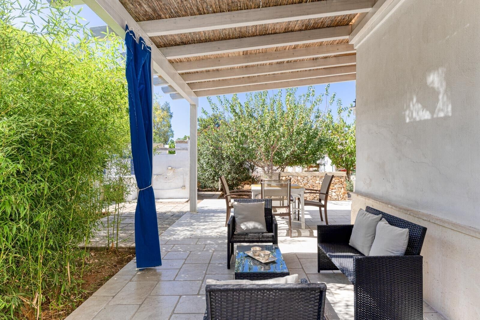 Un espace extérieur aménagé en terrasse, avec des meubles en osier, une table sur laquelle est posé un plateau, et un rideau bleu.