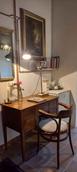 A wooden desk with a lamp, a chair, a vase with flowers, and a shelf with books.