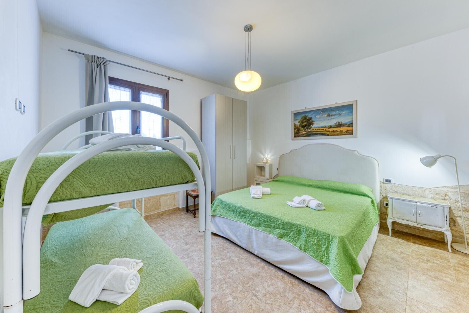 A hotel room with bunk beds, a bedside table with a lamp, a painting on the wall, and a window with curtains.