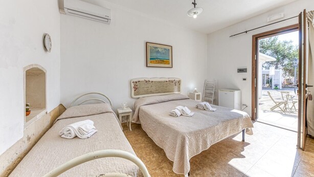 A room with two beds, a small table, a chair, an air conditioning unit, and a view of the outside. - Melograno Room (Porto Cesareo)