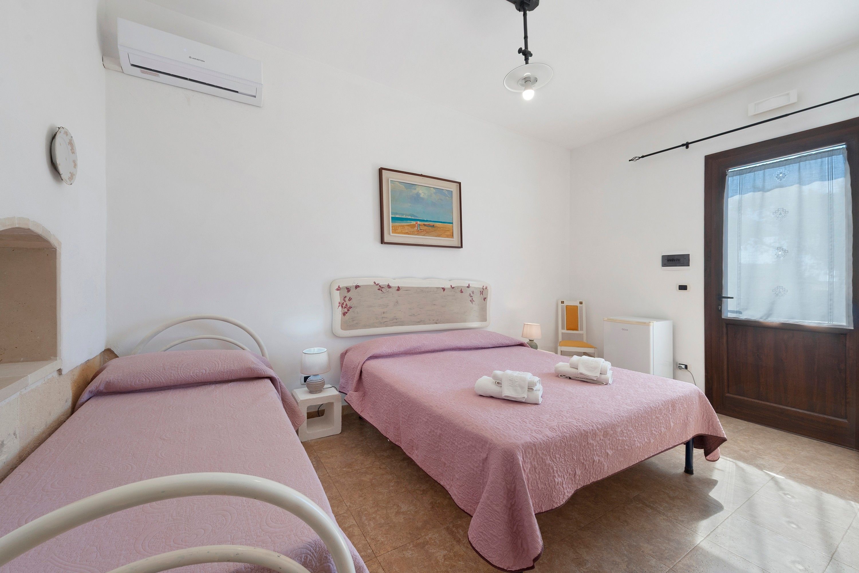 A hotel room with two beds, a painting on the wall, a window with curtains, and a door.