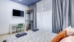 A bedroom with a bed, a desk, a chair, a TV, and a window with curtains.