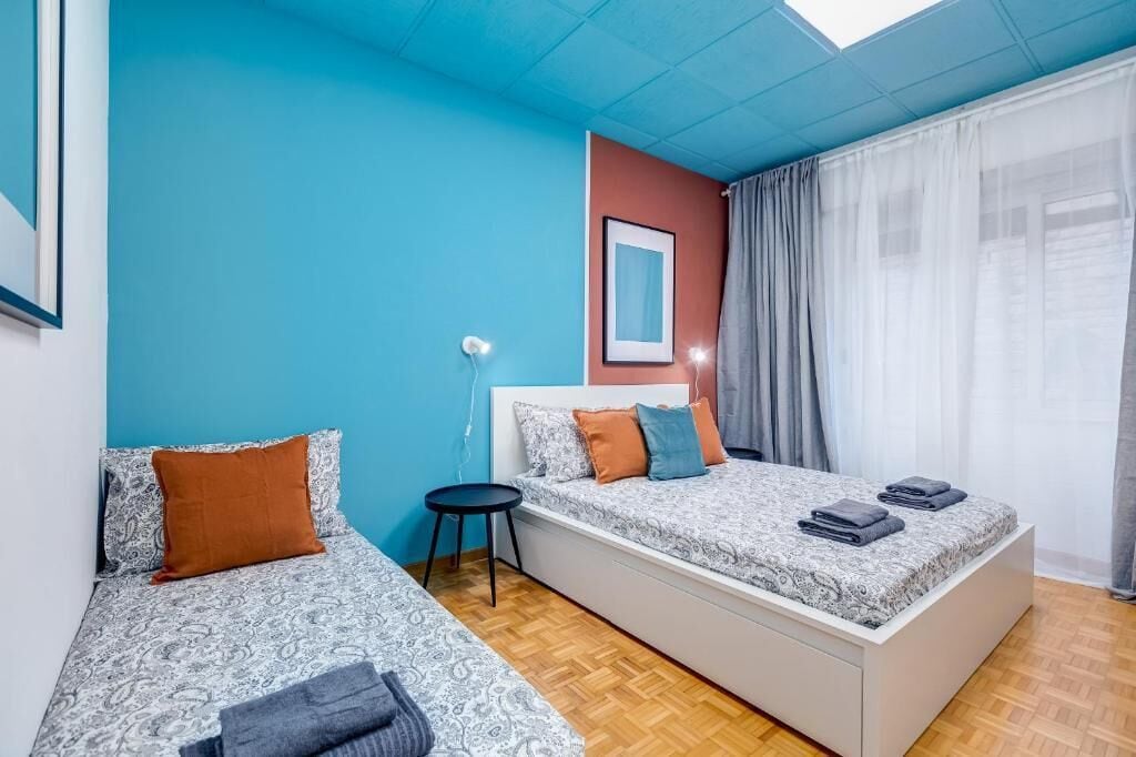 Una habitación con dos camas, una mesita, una lámpara en la pared y una ventana con cortinas.