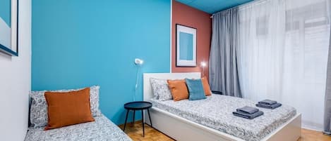 A room with two beds, a small table, a wall-mounted light, and a window with curtains.