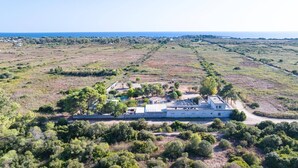 A large, rectangular building with a flat roof, surrounded by a natural landscape with sparse vegetation and a distant view of the sea. - Corbezzolo Room – Ground Floor, 4 Beds, Air Conditioning, Private Bath (Porto Cesareo)