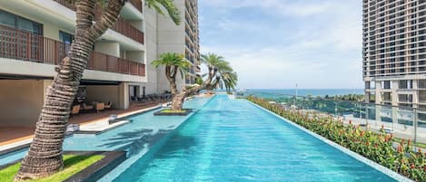 A long swimming pool with a clear view of the ocean, surrounded by palm trees and modern buildings.