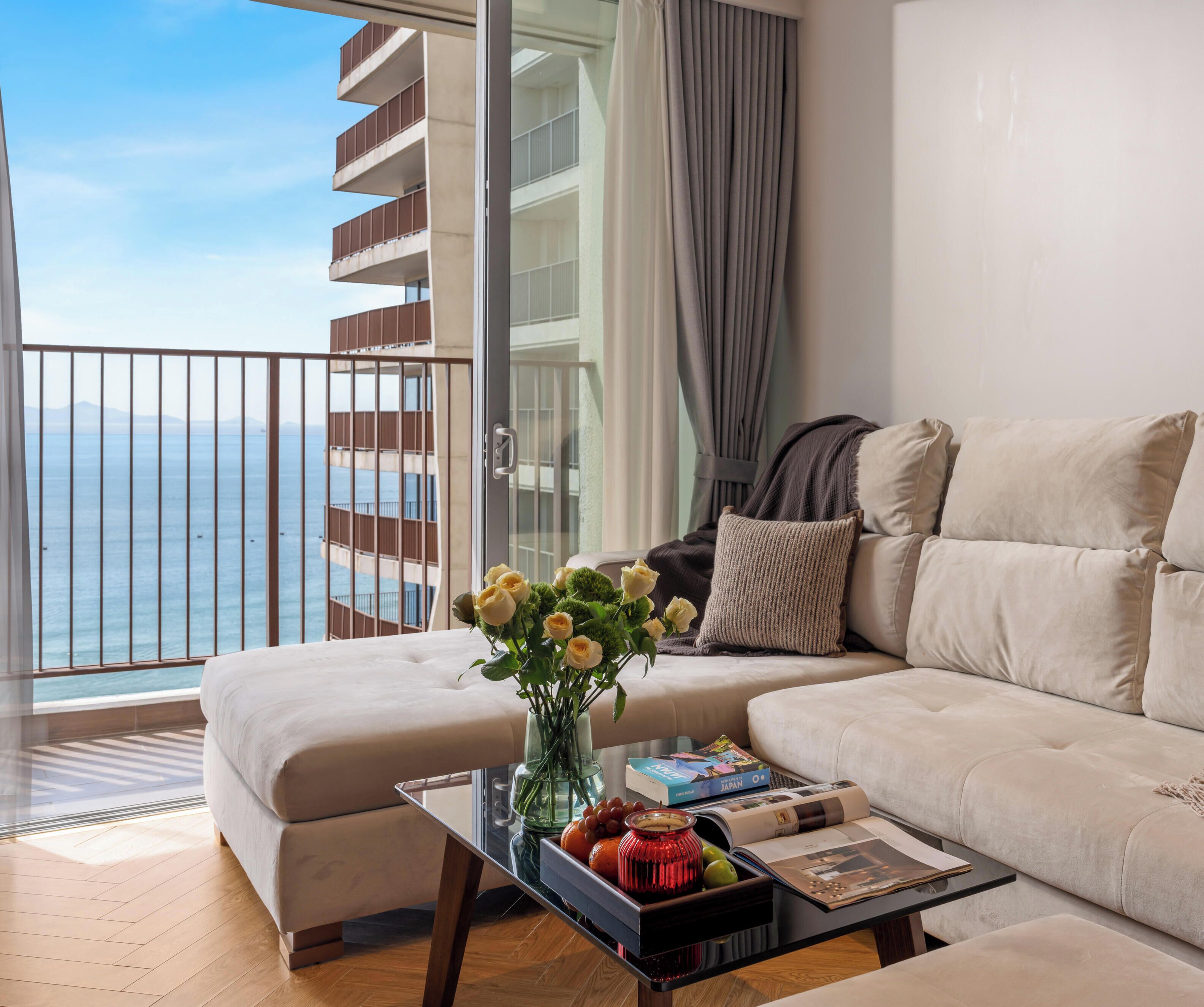 A modern living room with a sofa, a coffee table with a vase of flowers, and a view of the sea through a balcony.