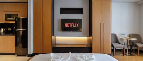 A hotel room with a bed, a TV displaying 'NETFLIX', a kitchenette, and a seating area with two chairs and a table.