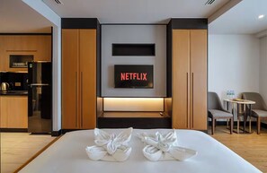 Habitación de hotel con cama, televisor mostrando 'NETFLIX', kitchenette y una zona de estar con dos sillas y una mesa.