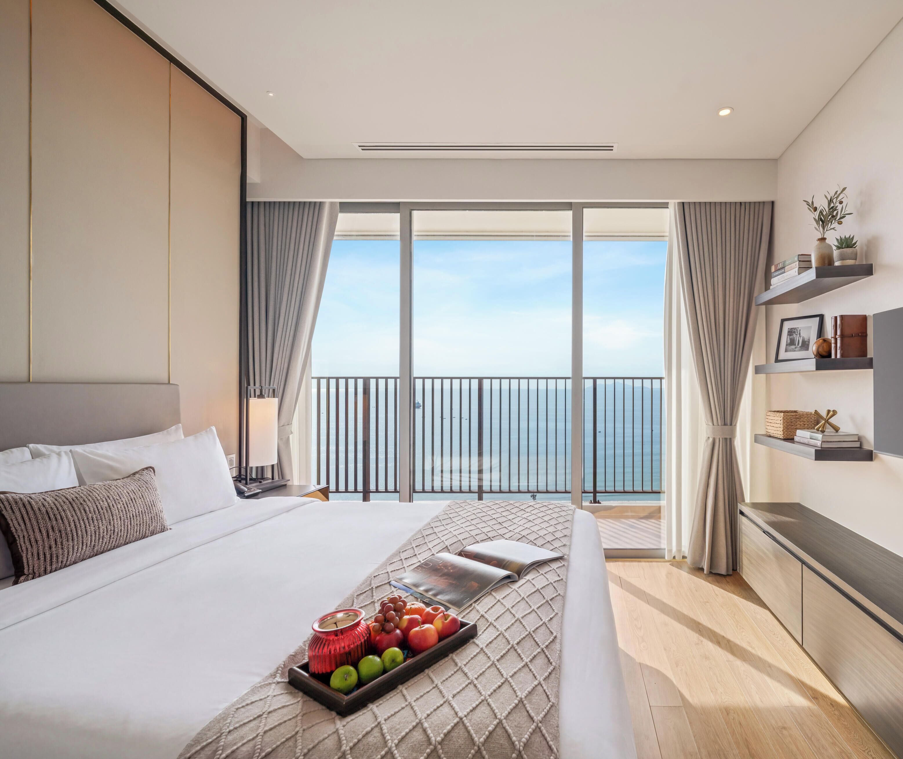 A hotel room with a large bed, a bedside table, a tray of fruit, a window with a view of the sea, and a balcony with a railing.
