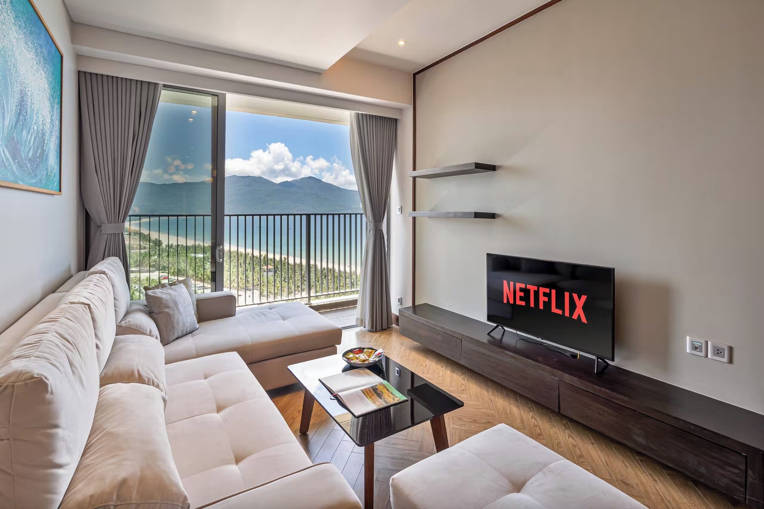 A modern living room with a large TV displaying 'NETFLIX', a sofa, a coffee table, and a balcony with a view of the sea.