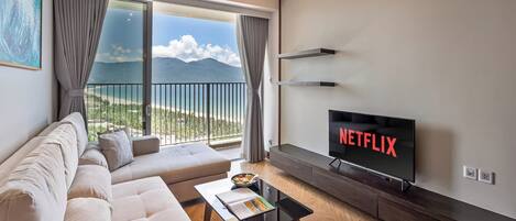 A modern living room with a large TV displaying 'NETFLIX', a sofa, a coffee table, and a balcony with a view of the sea.