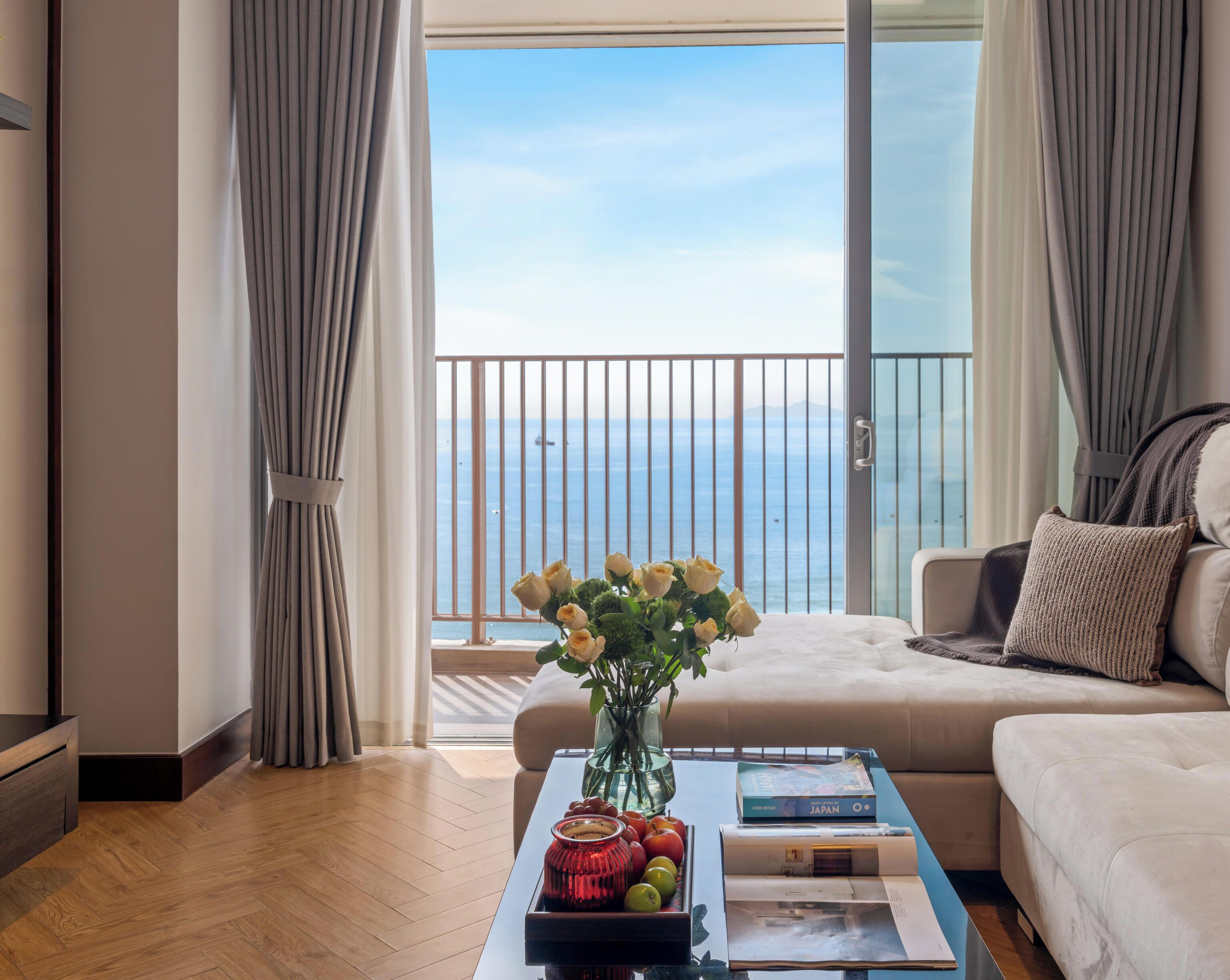 A modern living room with a view of the sea, a sofa, a coffee table with books and fruit, and a vase with flowers.