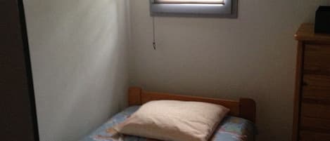 Una habitación pequeña con cama individual, cabecera de madera, ventana con persianas y cómoda de madera.