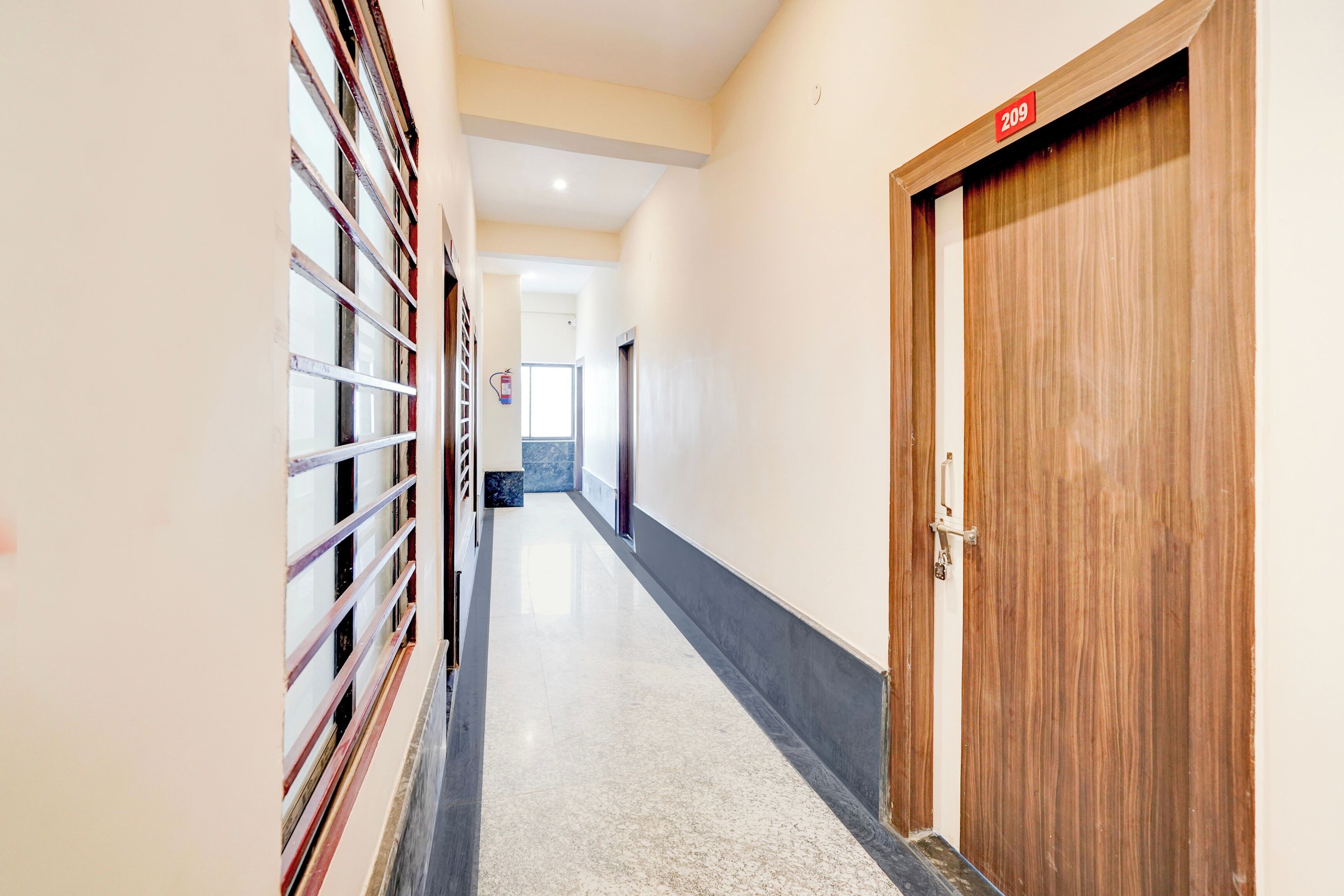 Un pasillo con una puerta de madera marcada con el número 209, un extintor en la pared y una ventana con barrotes.