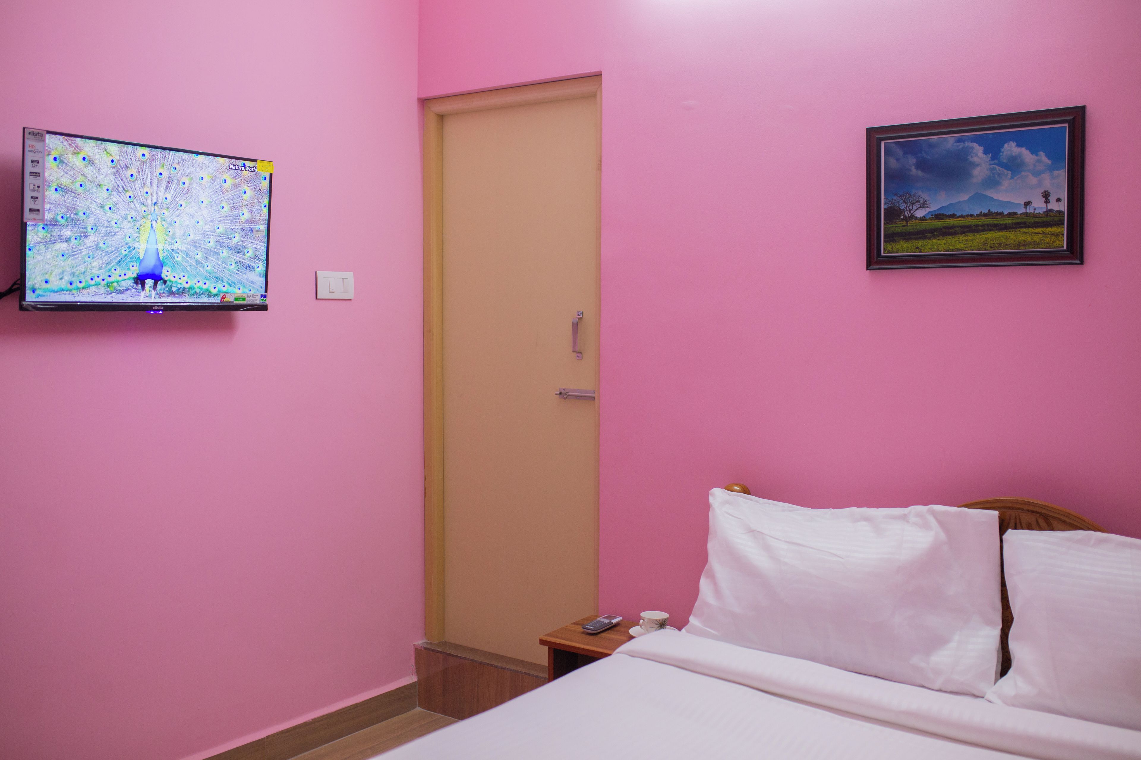 Una habitación con cama, un mesita de noche con una taza y un control remoto, un televisor en la pared y una puerta.