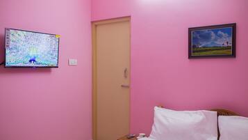 Una habitación con cama, un mesita de noche con una taza y un control remoto, un televisor en la pared y una puerta.