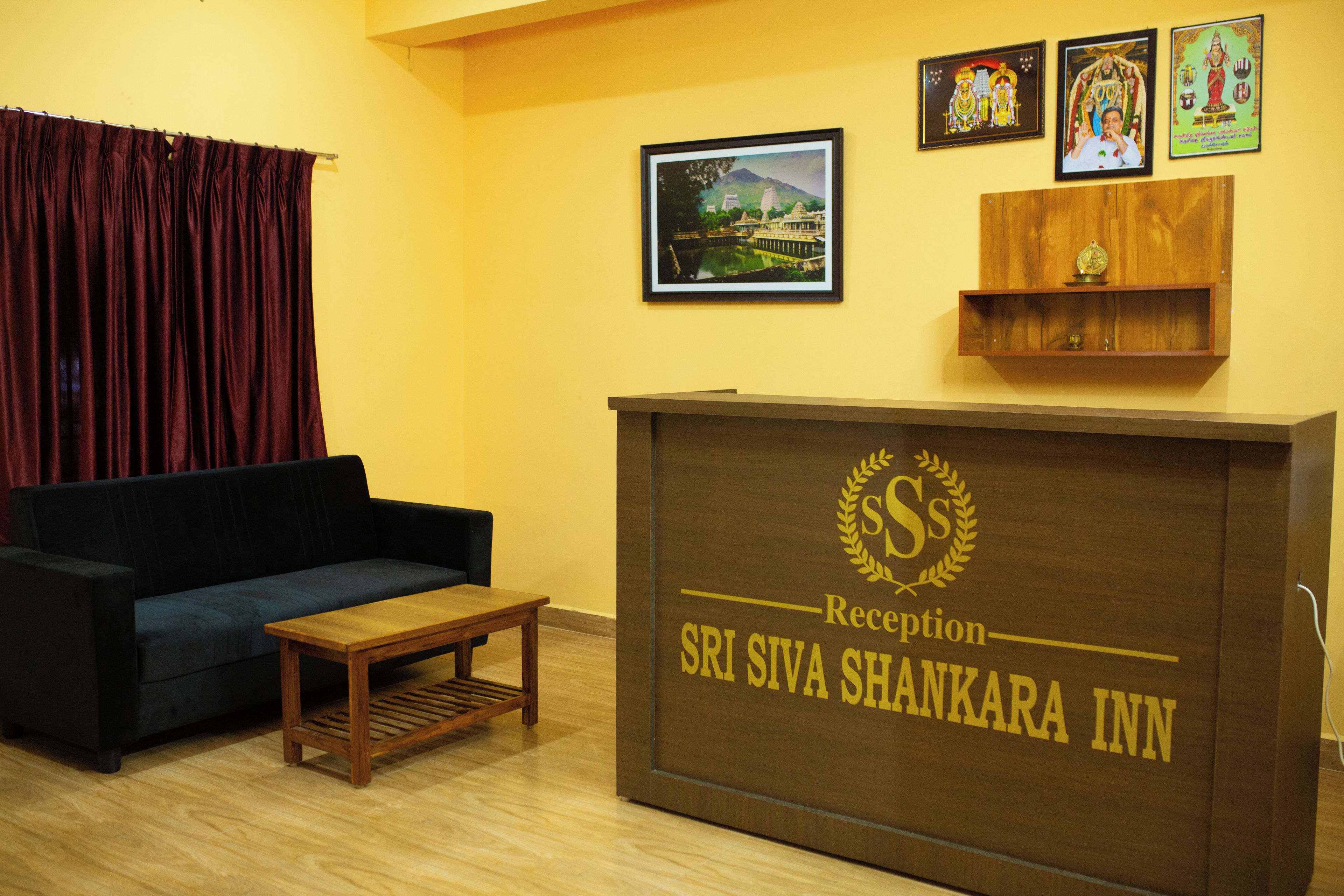 Un mostrador de recepción con el nombre 'SRISIVA SHANKARA INN' y una zona de recepción con un sofá, una pequeña mesa y cuadros enmarcados en la pared.