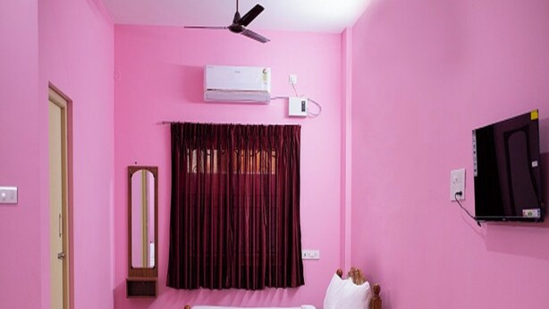 Een kamer met roze wanden, een bed, een plafondventilator, een airconditioningapparaat, een televisie en een klein tafeltje met flessen.