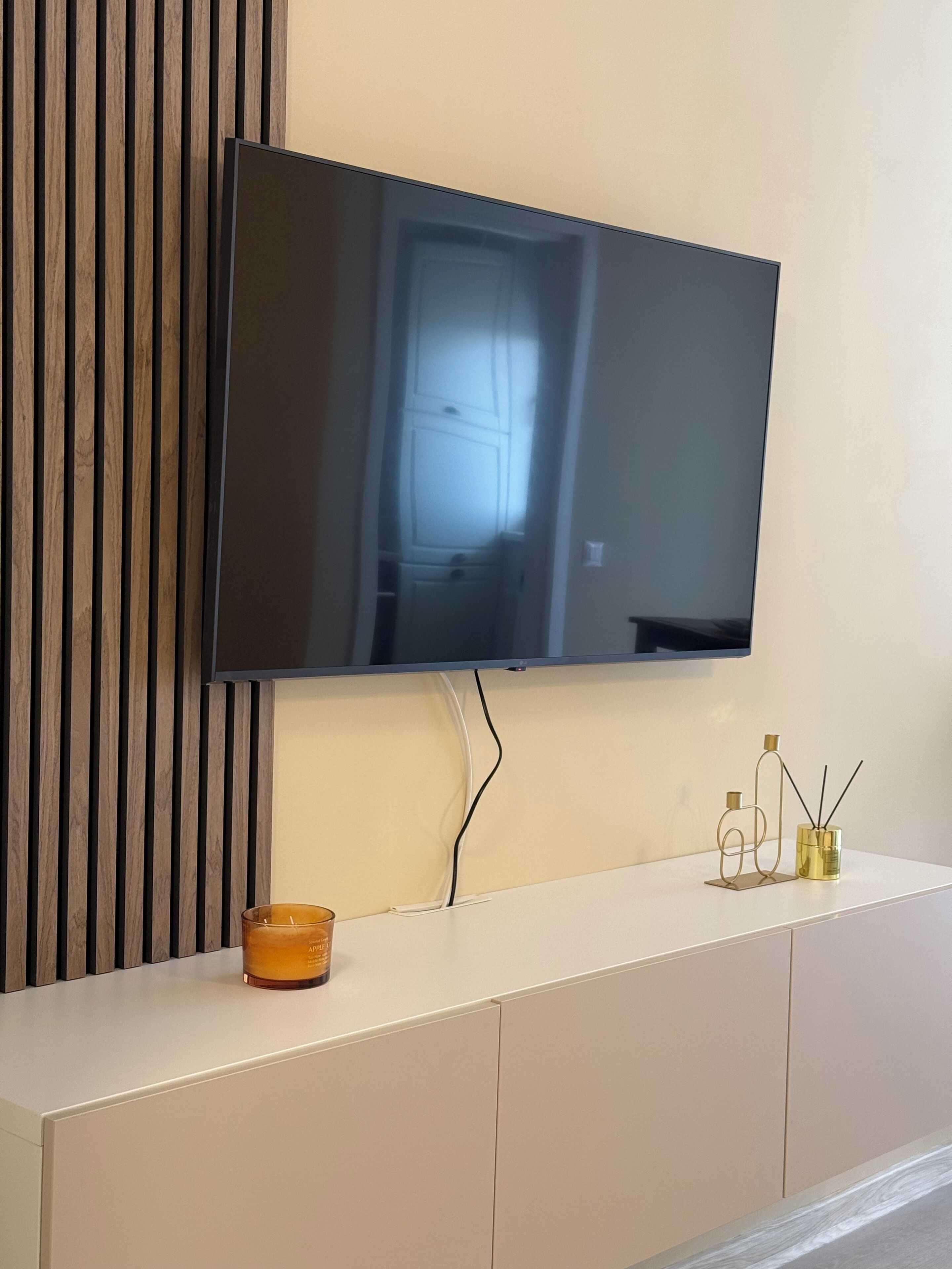 A flat-screen TV mounted on a wall above a white cabinet with a candle and reed diffuser on it.