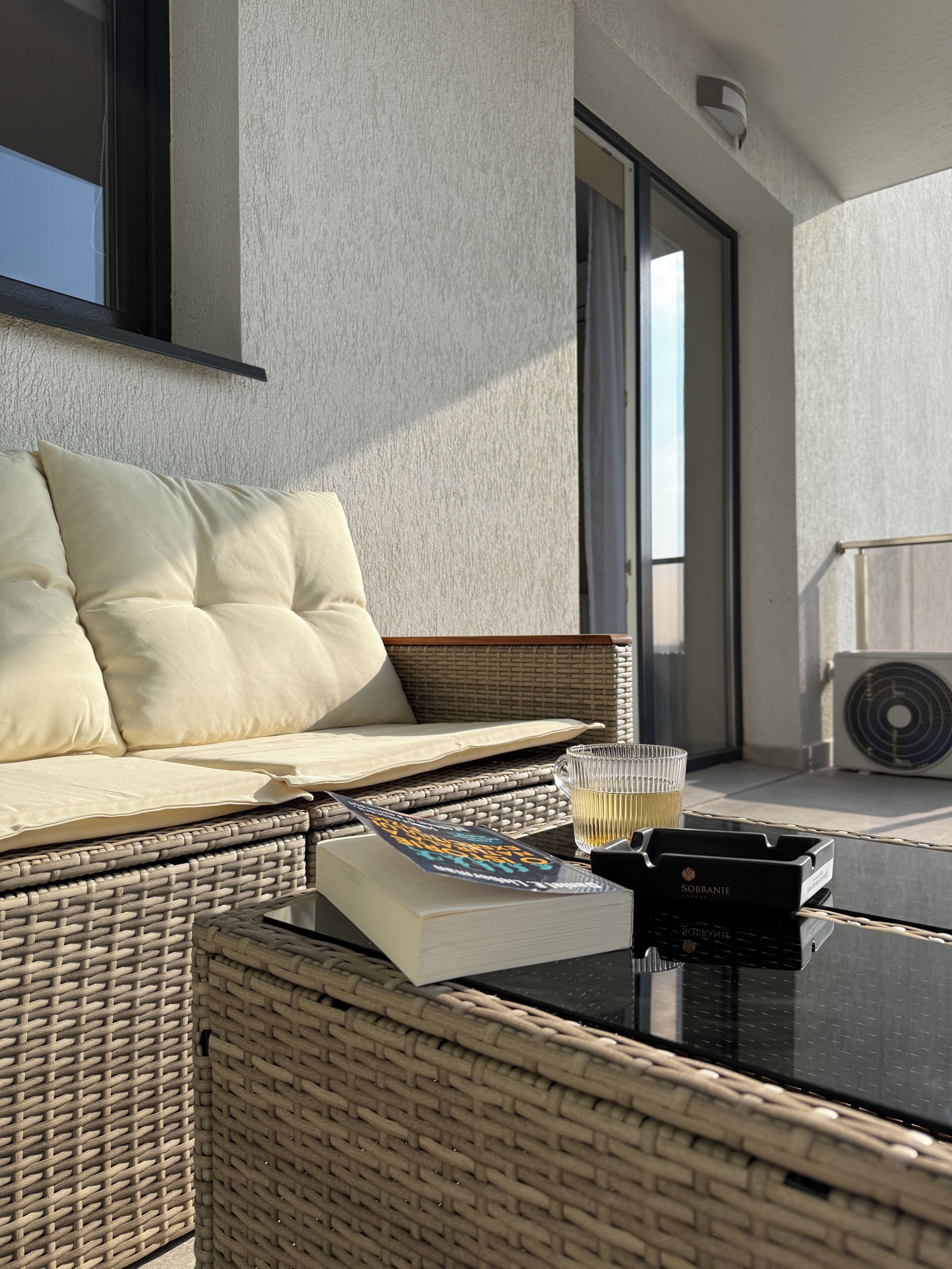 A balcony with a wicker sofa, a glass table, a book, a glass, and a black ashtray.