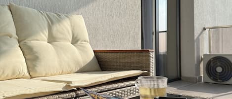 A balcony with a wicker sofa, a glass table, a book, a glass, and a black ashtray.