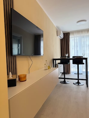 Une chambre d’hôtel moderne dotée d’un grand téléviseur à écran plat fixé au mur, d’un petit coin bar avec des tabourets et d’une fenêtre ornée de rideaux légers.