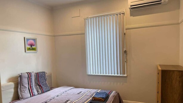 Un dormitorio con una cama, una ventana con persianas, un equipo de aire acondicionado y un armario de madera.