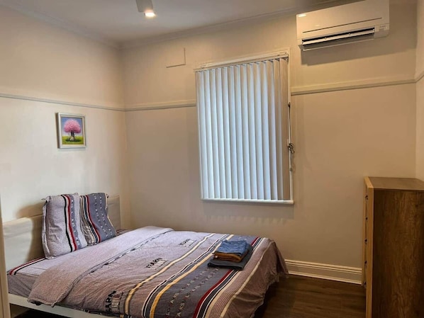 A bedroom with a bed, a window with blinds, an air conditioning unit, and a wooden wardrobe.