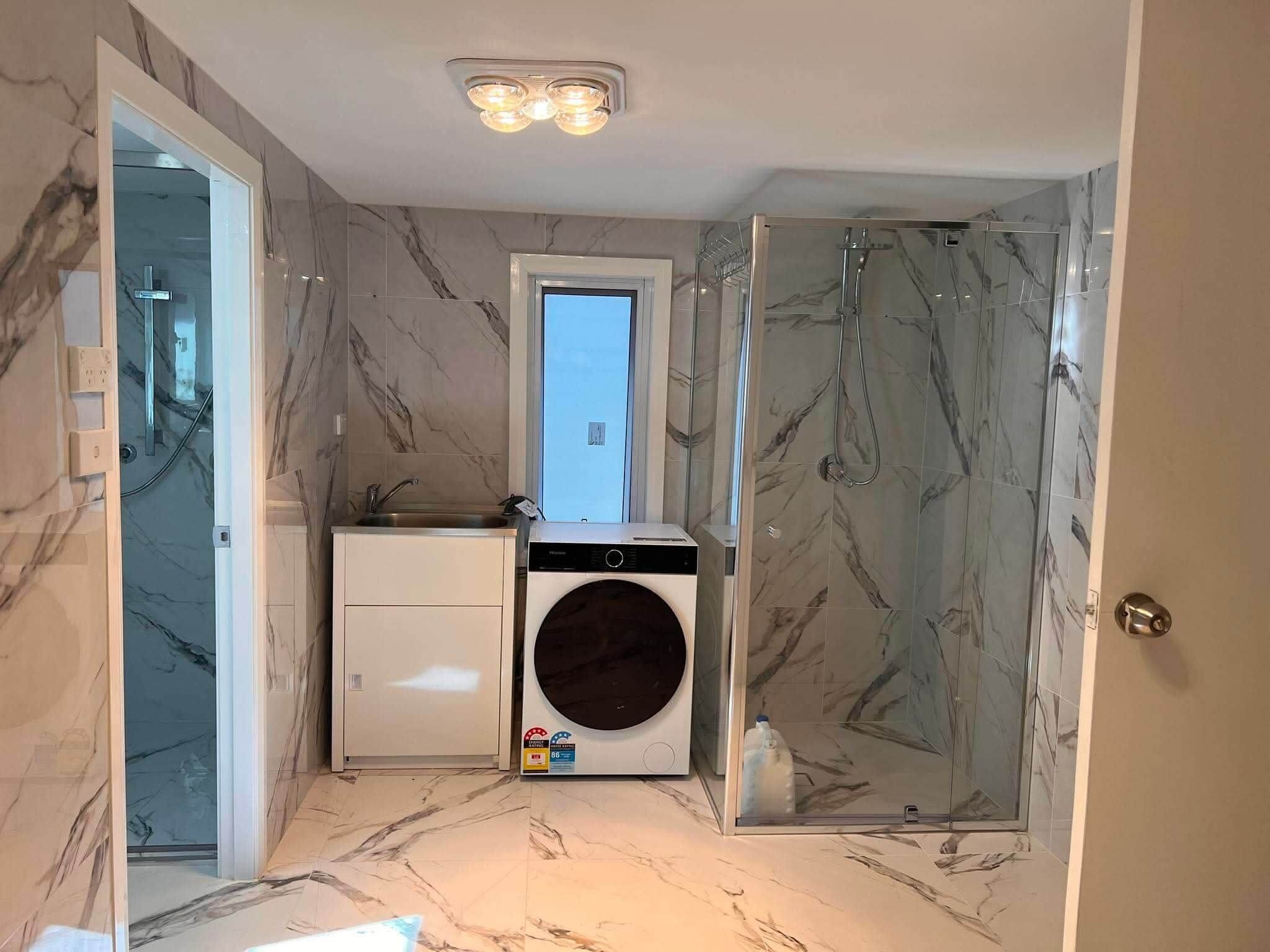 A compact bathroom with a glass shower enclosure, a washing machine, and a sink with a marble backsplash.