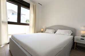 A bedroom with a large bed, two bedside tables, a window with curtains, and a view of buildings outside. - SUNNY HILLS dream apartment (Finestrat)