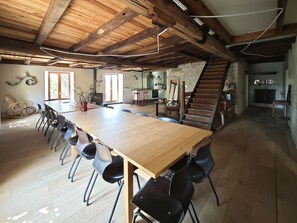 A spacious dining area with a long wooden table and black chairs, a kitchen in the background, and a wooden staircase.