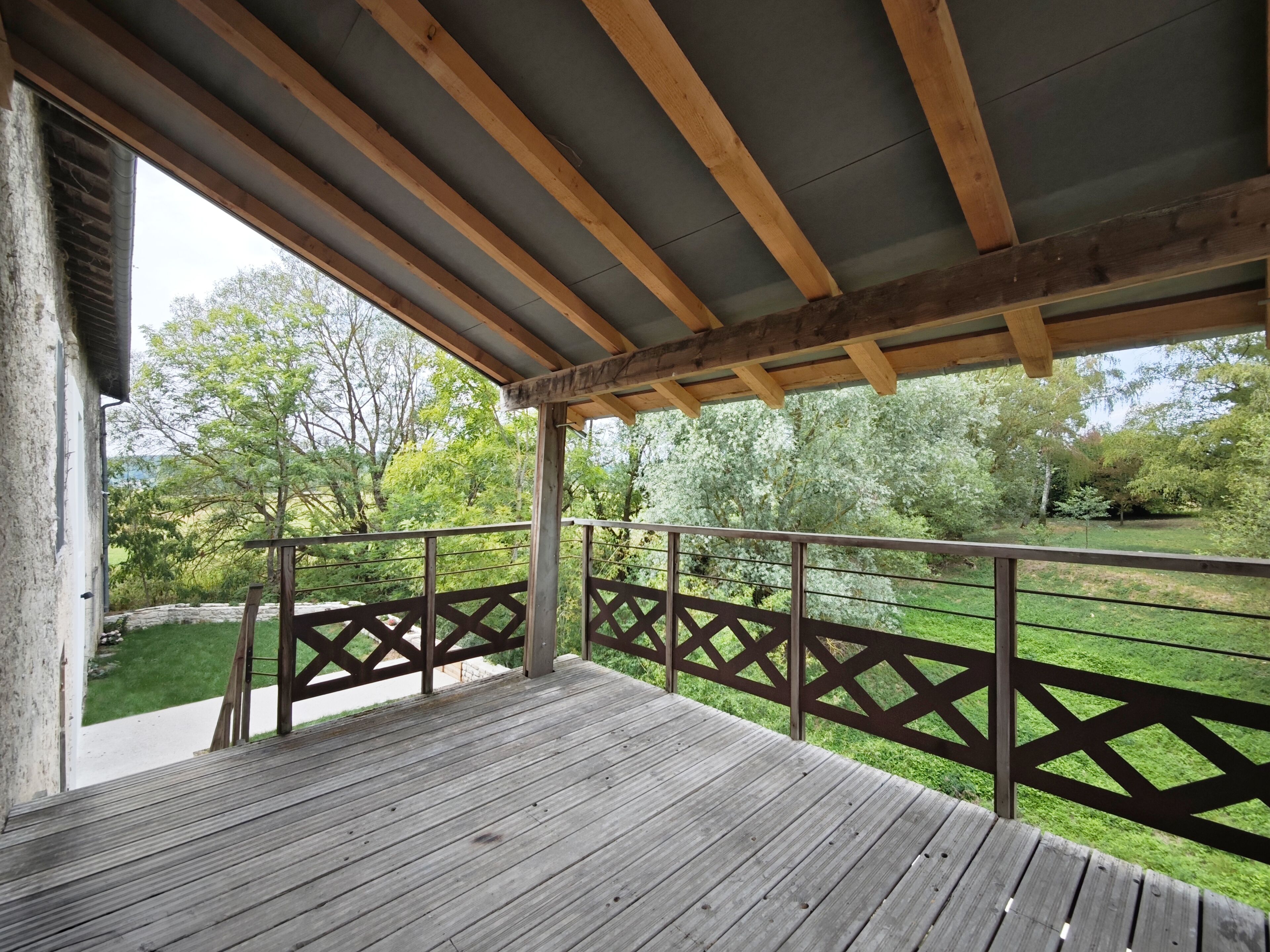 Une terrasse en bois avec une rambarde en métal, donnant sur un espace verdoyant planté d’arbres.