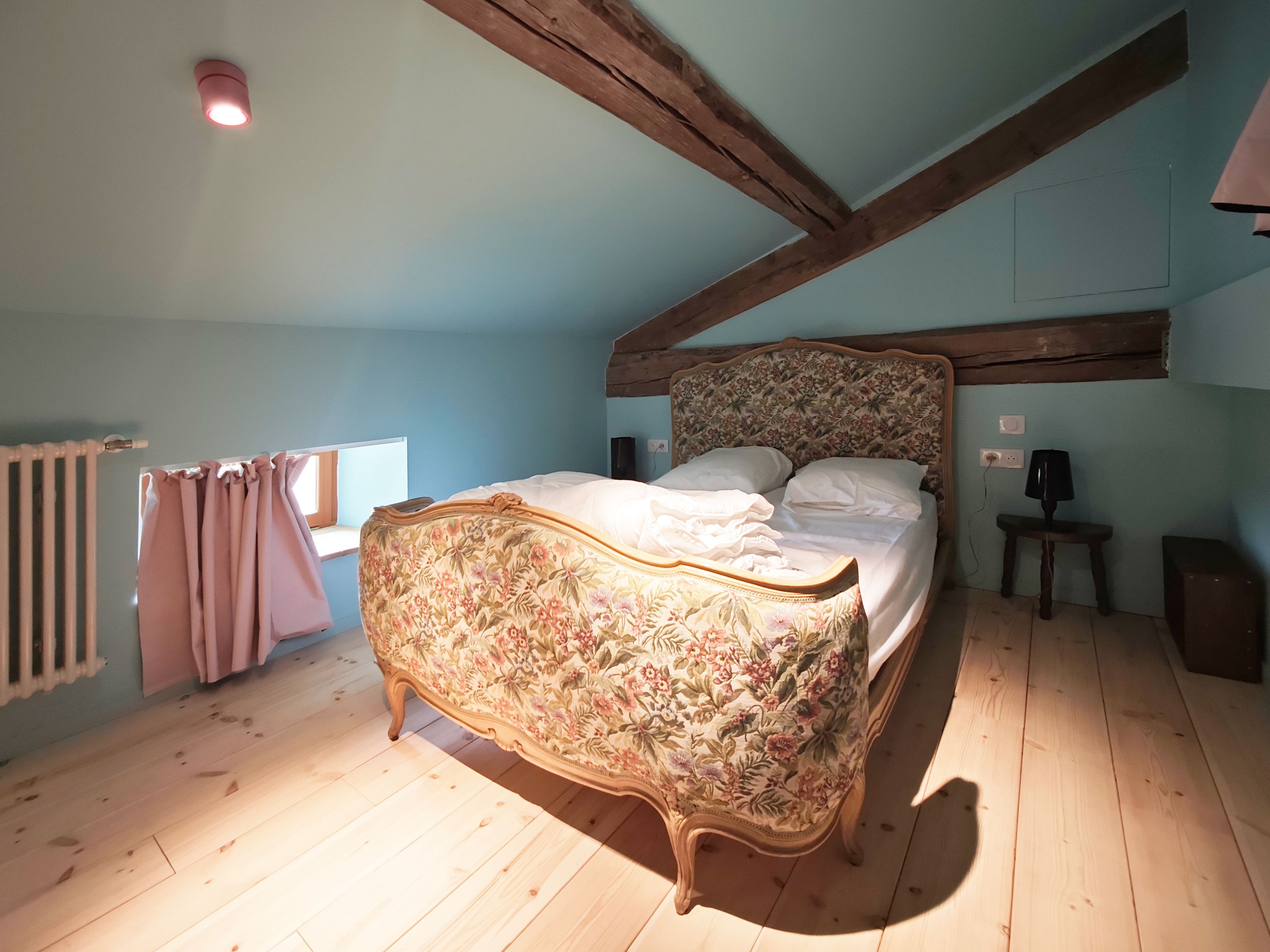 A bedroom with a floral-patterned bed, wooden beams, a radiator, and a window with curtains.