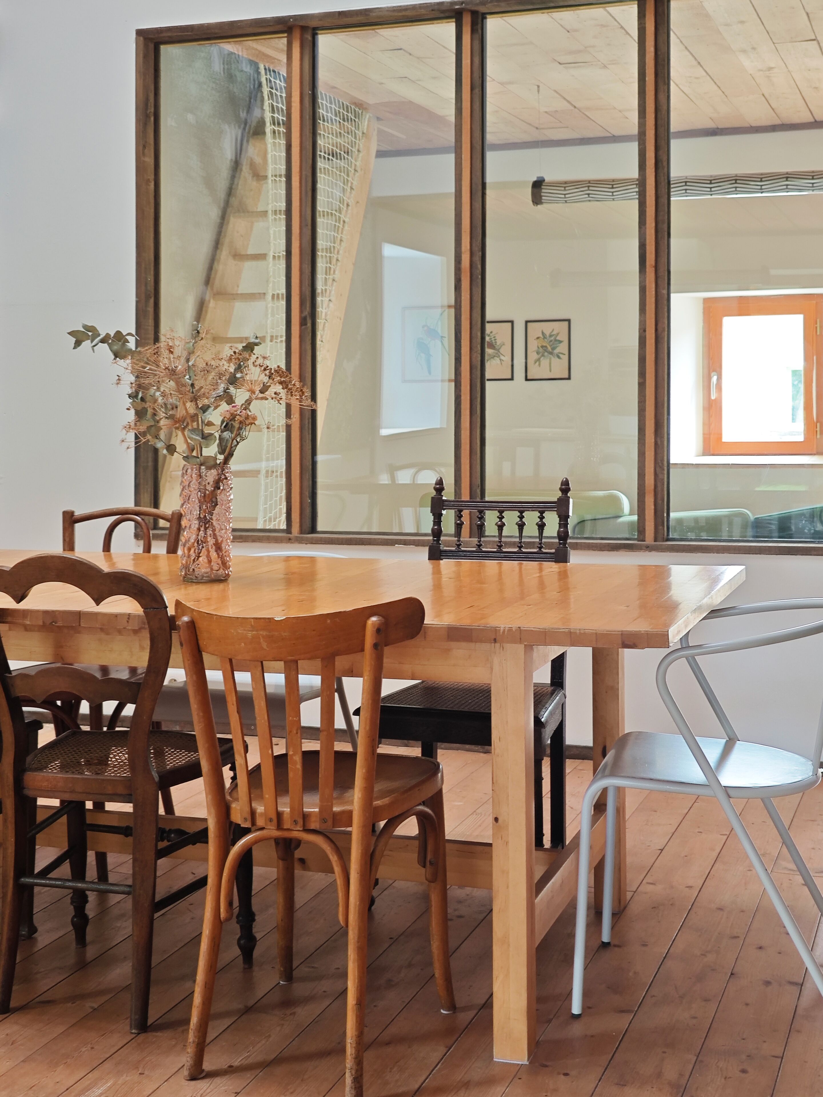 Une table à manger en bois, avec des chaises, un vase contenant des fleurs séchées, et une vue sur un escalier et une fenêtre.
