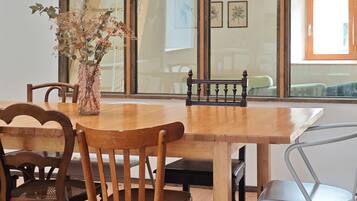 Une table à manger en bois, avec des chaises, un vase contenant des fleurs séchées, et une vue sur un escalier et une fenêtre.