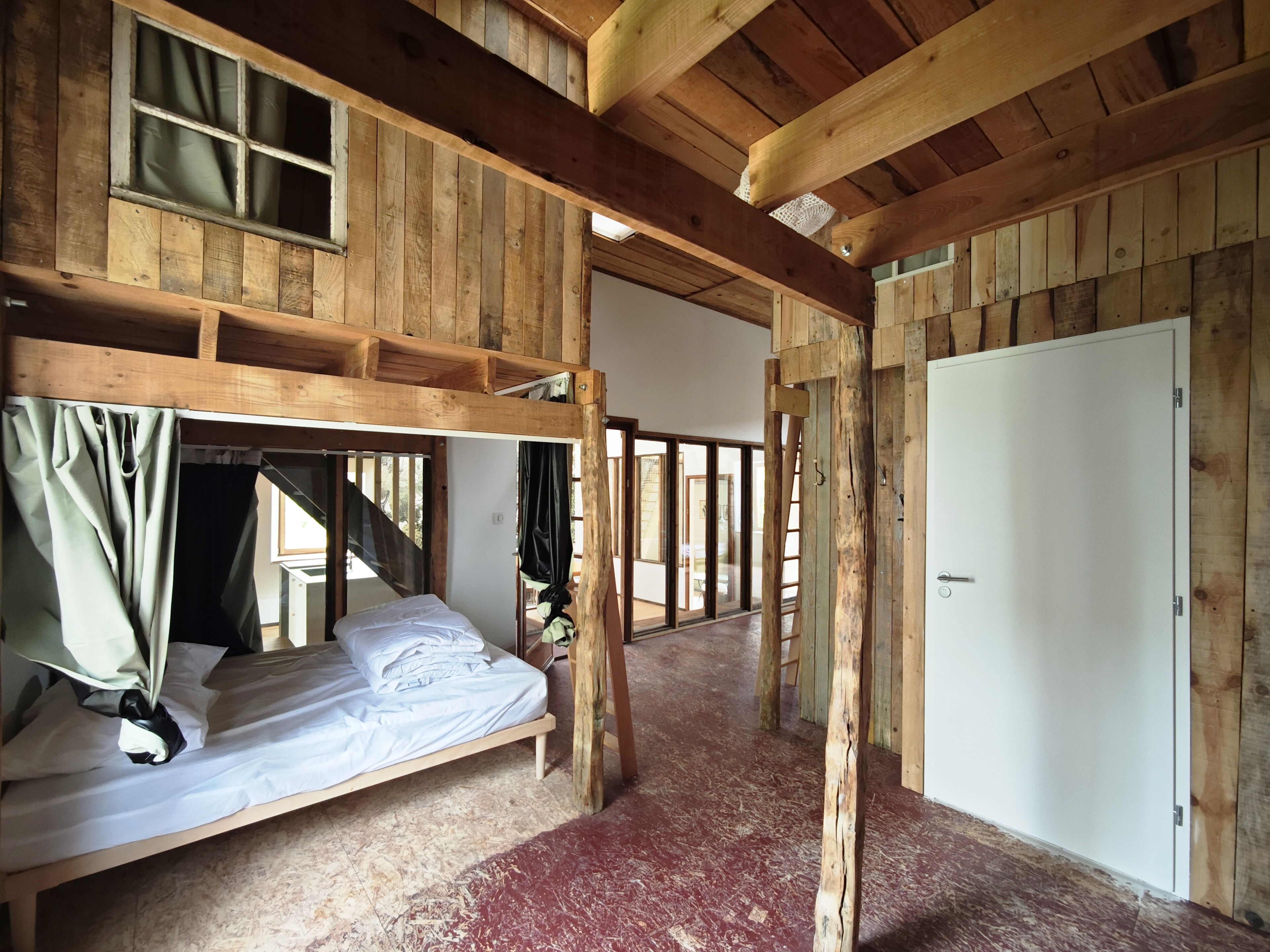 Une pièce rustique en bois, avec un lit superposé, une porte blanche et une fenêtre avec des rideaux.