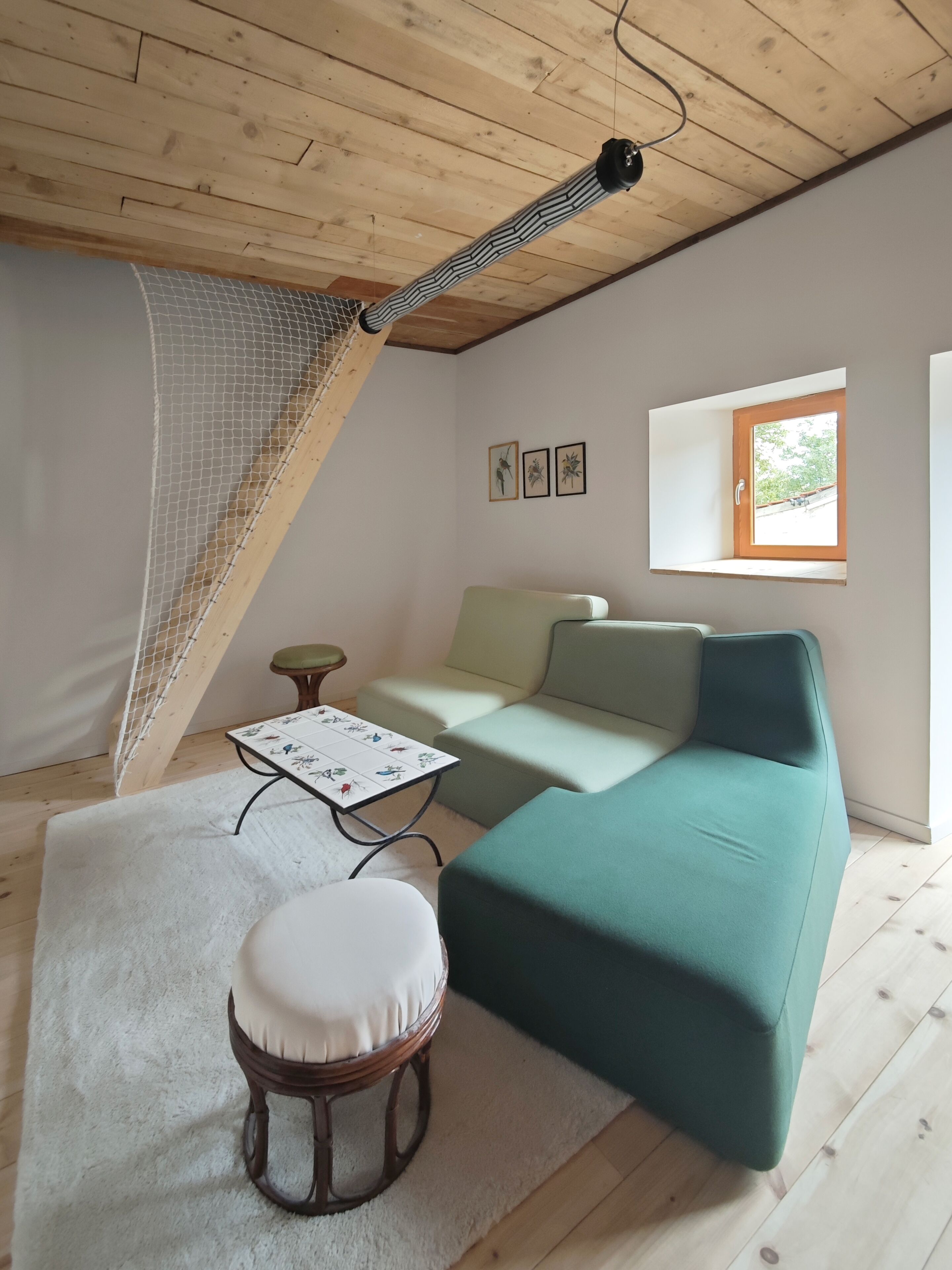 A cozy room with a wooden ceiling, a green sofa, a white cushioned chair, a small table with a floral tablecloth, and a window with a view of greenery.