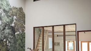 A dining area with a wooden table and chairs, a large window with a view, and a wall with a green leafy mural. - Domaine du Moulin Français - Outdoors (Champougny)