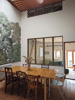 Een eethoek met een houten tafel en stoelen, een groot raam met uitzicht, en een muur met een groene, bladerige muurschildering.