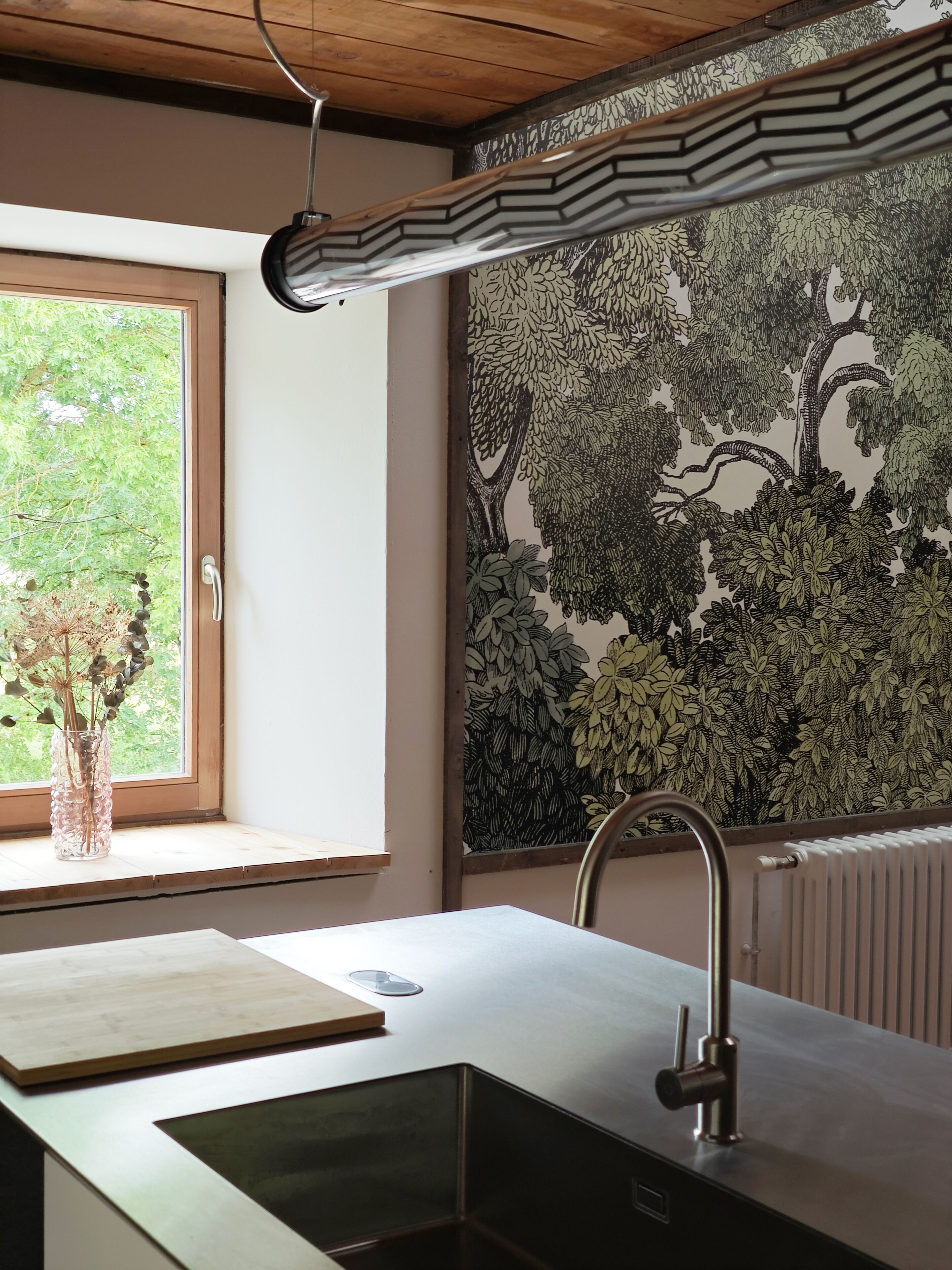 A modern kitchen with a wooden ceiling, a window with a view of trees, a radiator, and a sink with a faucet.