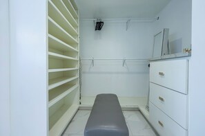 Et aflangt walk-in closet med hvide hylder, en bænk og et lille skab.