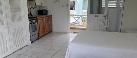 Une chambre comprenant un lit, une kitchenette équipée d’une cuisinière et d’un four à micro-ondes, une porte donnant sur un balcon et une fenêtre avec des stores.