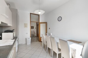 Een moderne keuken met witte kastjes, een magnetron en een spoelbak. Er is een eethoek met een tafel en stoelen, en een klok aan de muur.