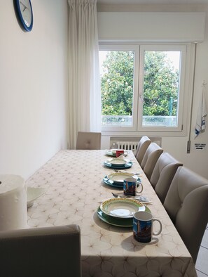 A dining area with a table set for four, a clock on the wall, and a window with a view of greenery. - Aaron Guest House - 4-Bedroom Apartment in Marghera (Venice)